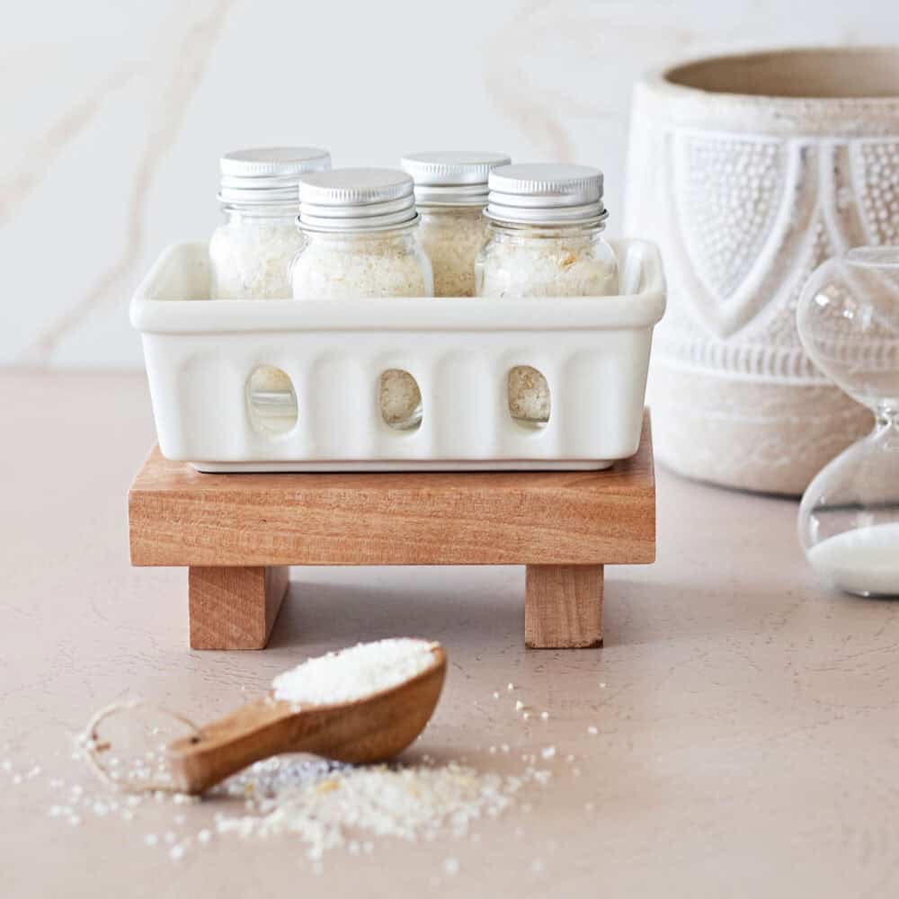 Jars of oatmeal bath ingredients in a white ceramic basket on a wood tray, behind a scoop of a oatmeal bath mix.