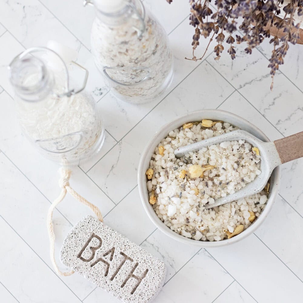 closeup of glass apothecary jars filling with a milk bath mixture from scoop and bowl under a canopy of dried lavender next to a pumice stone that says BATH