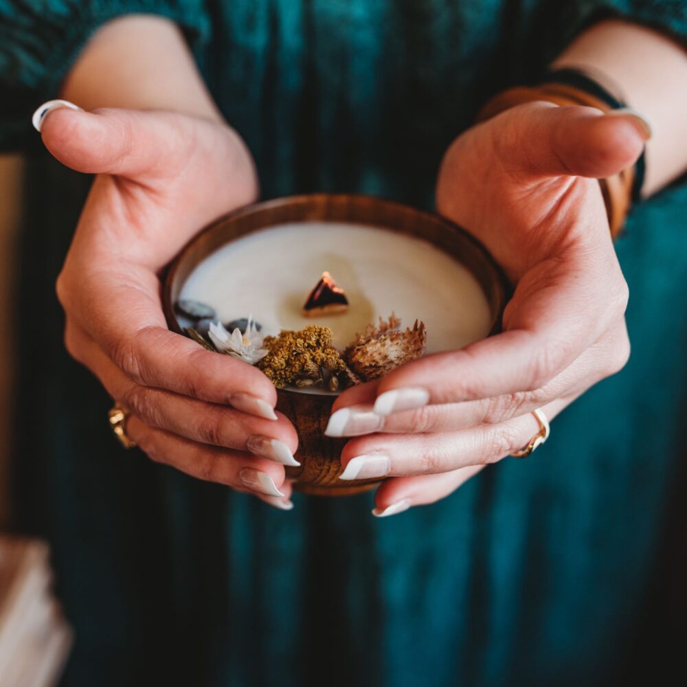 Kristy's hands holding a homemade botanical candle with a glowing wood wick.