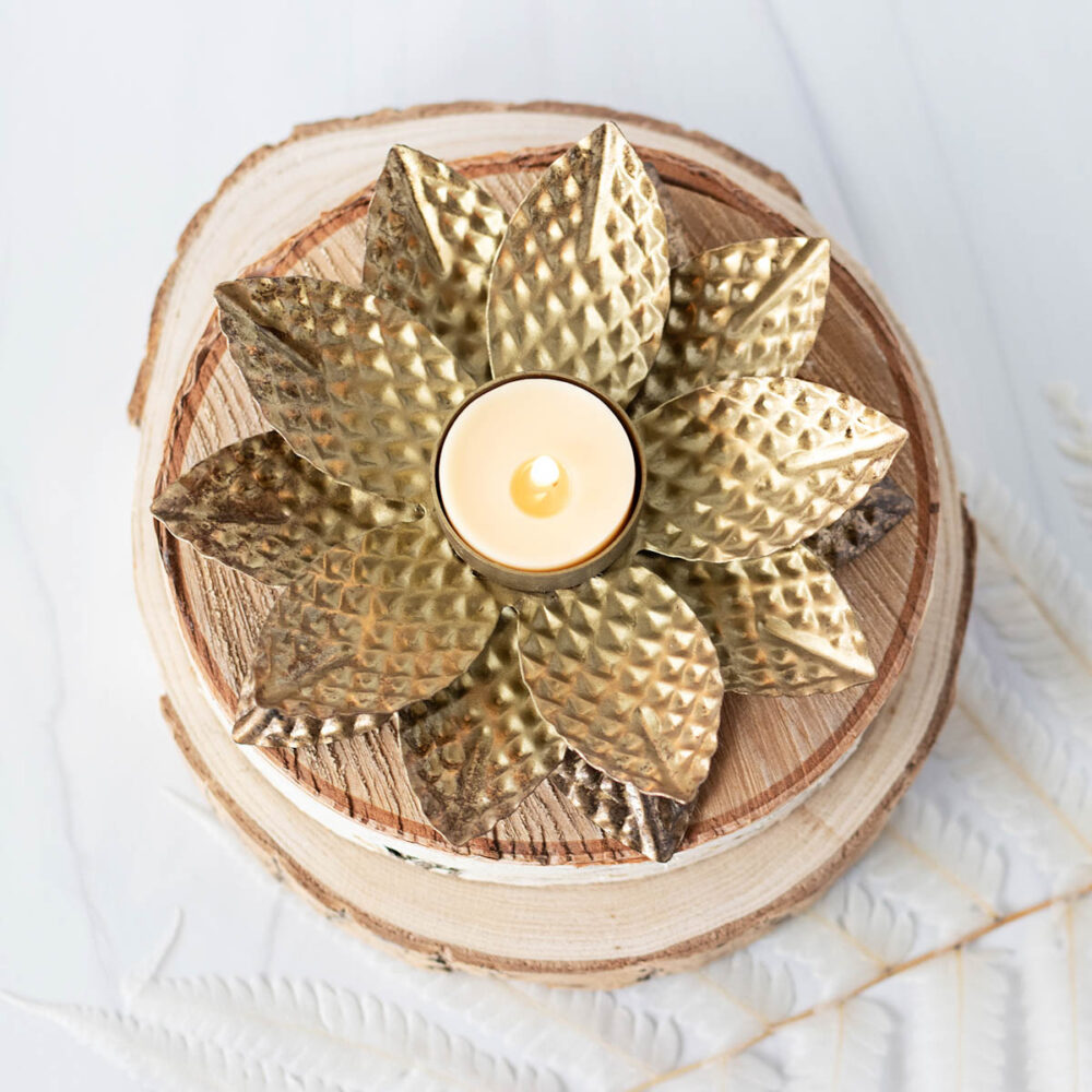 Glowing lit beeswax votive candle on rustic wood slices and a golden flower tea light holder.